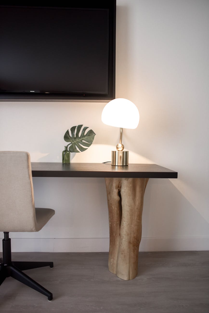 stainless steel base white shade table lamp on brown wooden desk near white painted wall with wall mounted flat screen t v